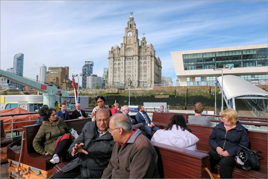 Two older Asian men in deep conversation on the top deck of a Mersey ferry boat with the Liver Building in the background