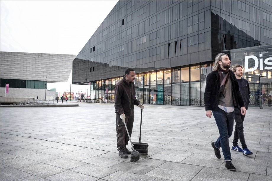 Two aloof young men ignore a coloured cleaner in a modern piazza.