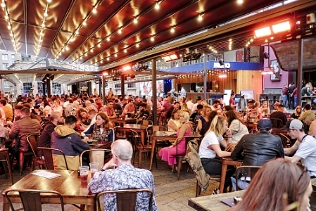 A girl looks towards a man sitting alone in a crowded bar.